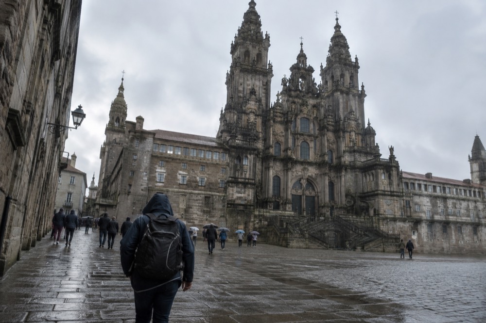 A pilgrim in a rain jacket walking through wet stone streets toward the cathedral in Santiago de Compostela