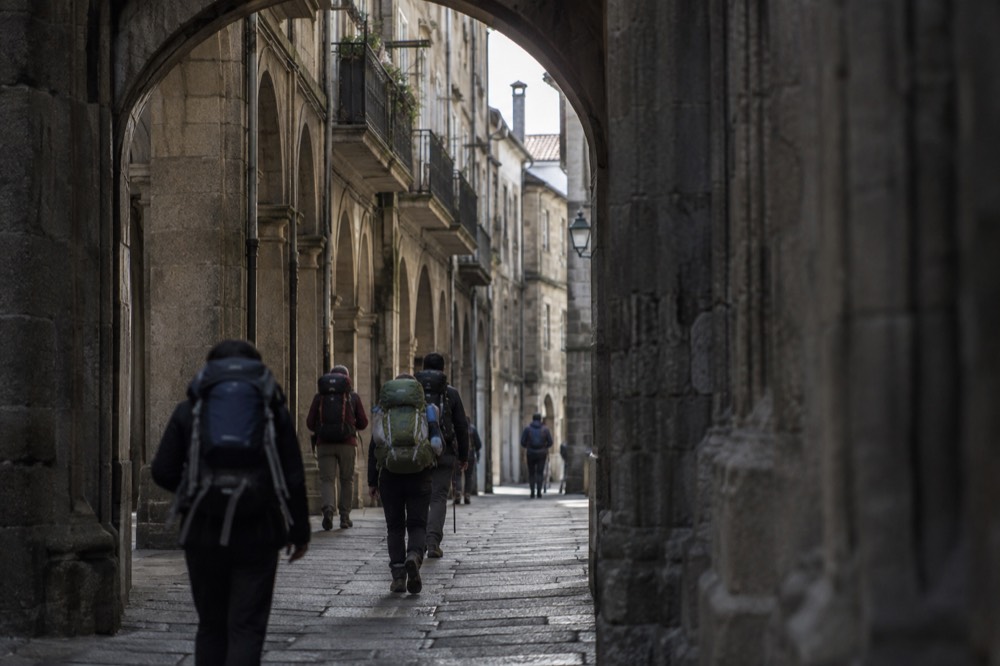 Pilgrims with backpacks walking through a stone archway into a Galician old town street