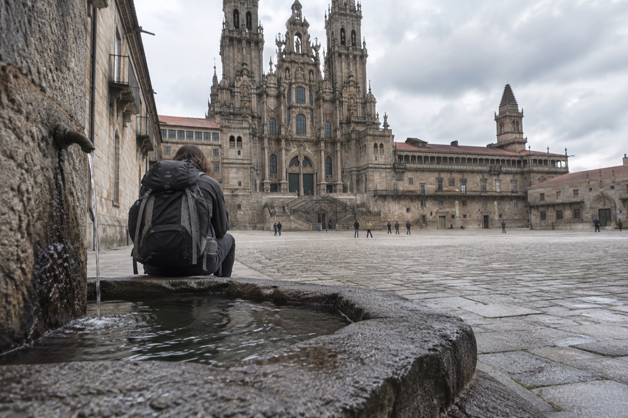 A pilgrim with a backpack resting beside a stone village fountain on a quiet Galician walking path