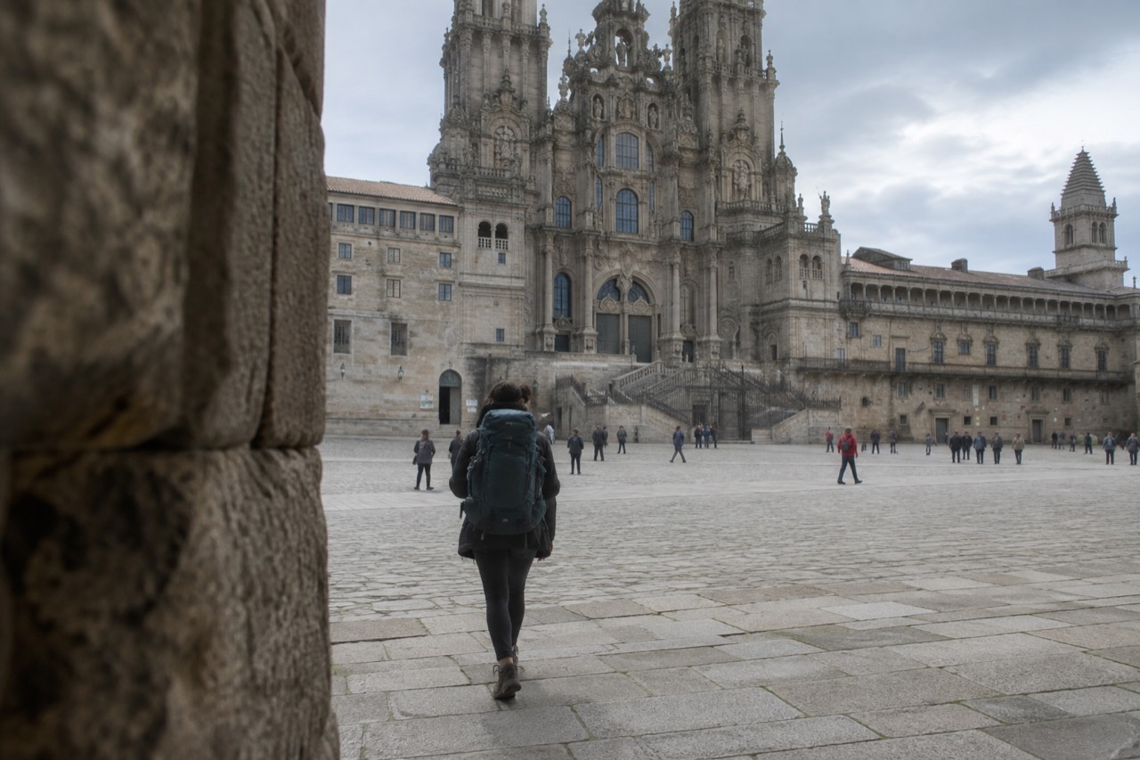 A pilgrim with a backpack walking a tree-lined rural path toward Santiago de Compostela