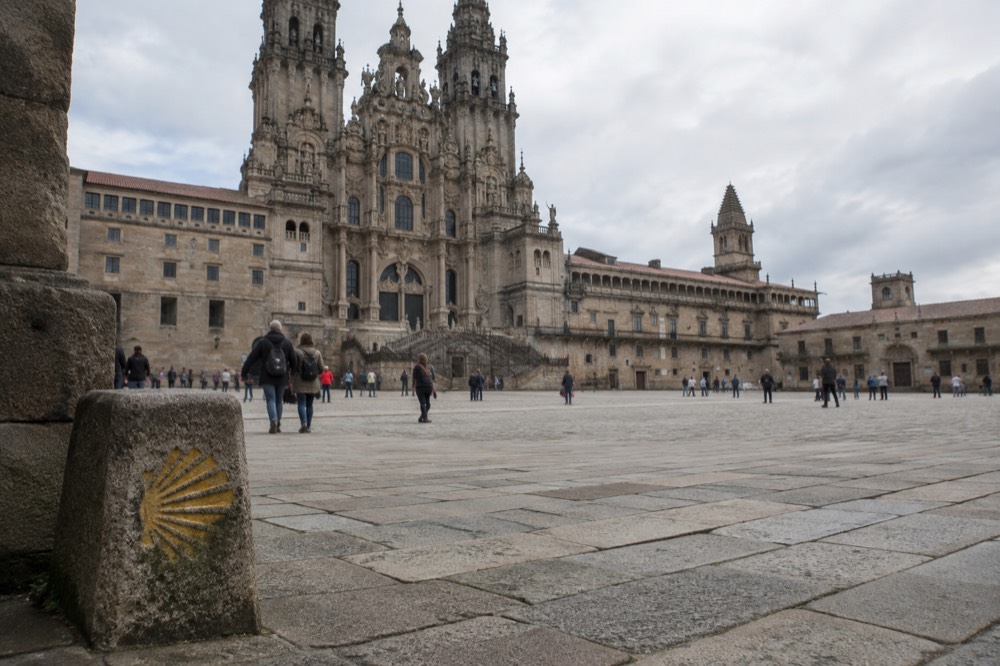 A yellow scallop-shell marker on a stone trail sign pointing a Camino walker toward Santiago de Compostela