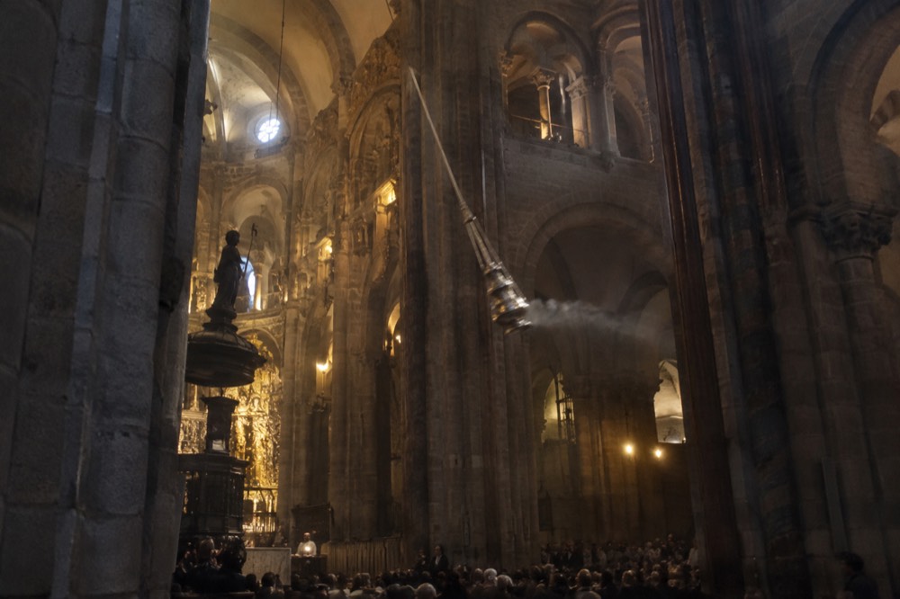 Pilgrims with backpacks and walking sticks gathered in a granite square beside a cathedral at golden hour