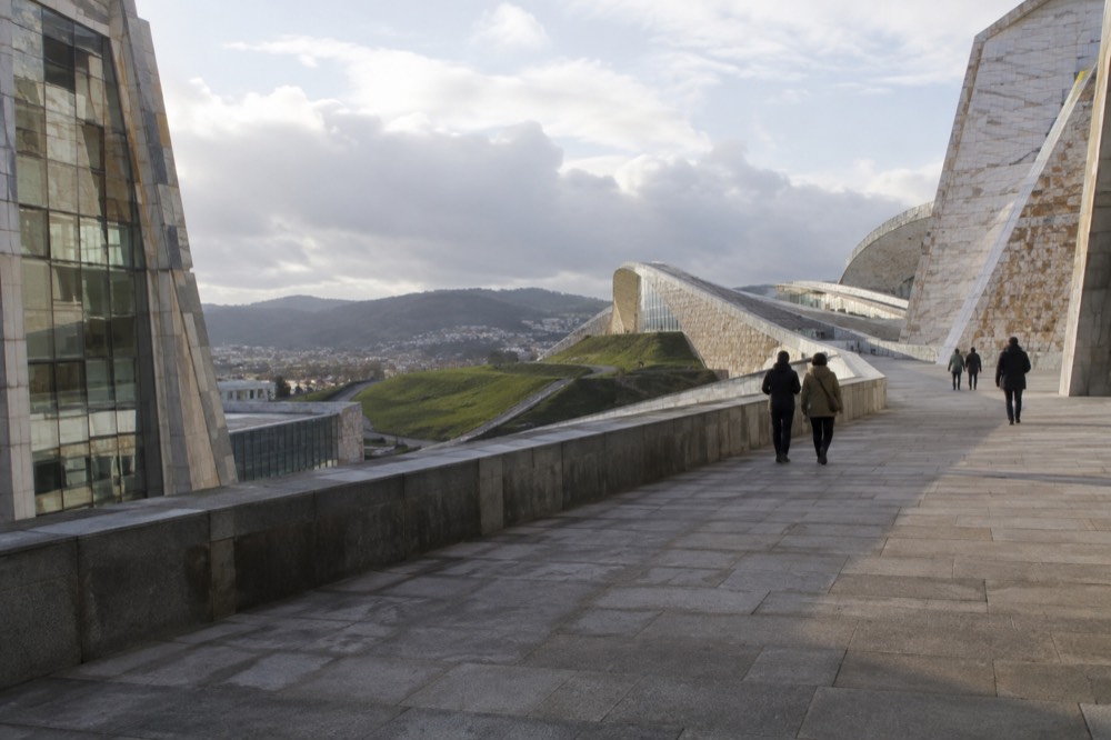 Pedestrians walking along a stone terrace path between modern buildings at the City of Culture