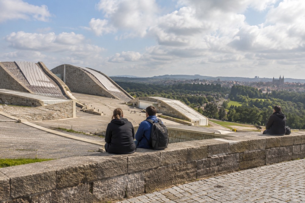 Travellers resting on a stone wall looking out over a green valley towards a historic city