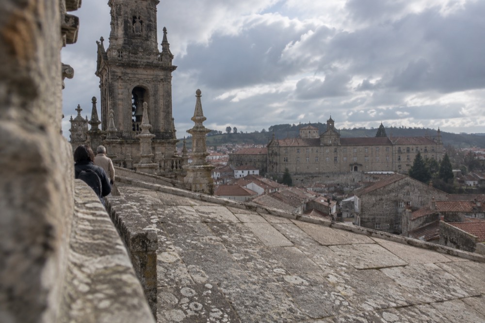 Visitors on a cathedral rooftop walkway looking across the tiled roofs toward the Parador building