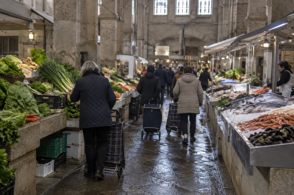 Shoppers with trolley bags moving between market stall rows under an awning
