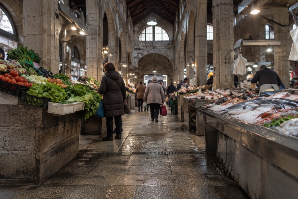 A covered market hall with produce and fish stalls and shoppers with baskets inside a granite building