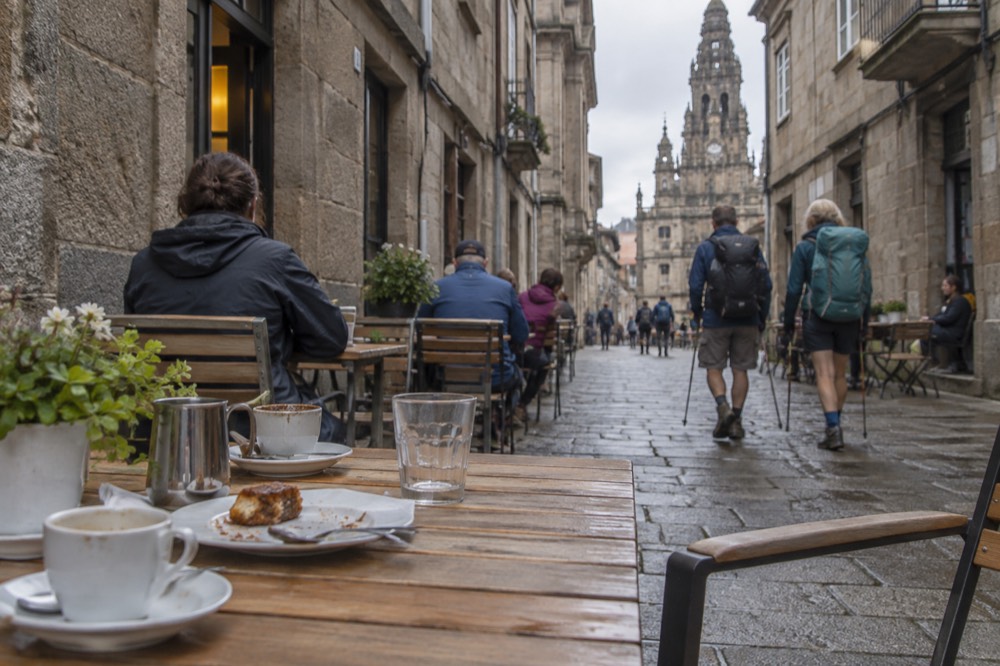 Pilgrims with backpacks and walking sticks resting at outdoor tables on a stone street beside a historic facade