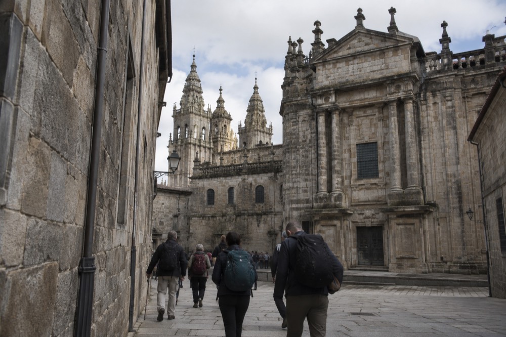 Travelers with backpacks walking through a narrow granite street toward the monastery church towers in Santiago de Compostela