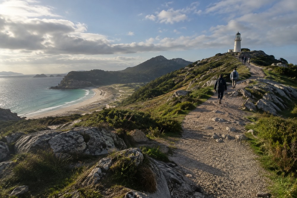 People walking a gravel path toward a lighthouse on a low green island summit