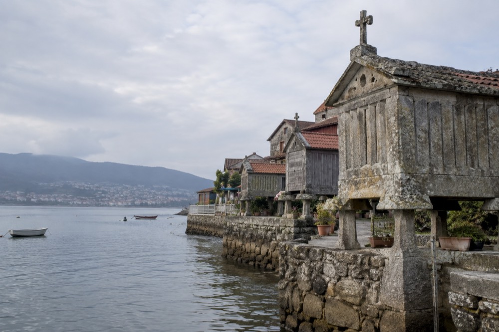 A close view of a stone hórreo on granite pilings at the water's edge, its gable topped with a small carved cross