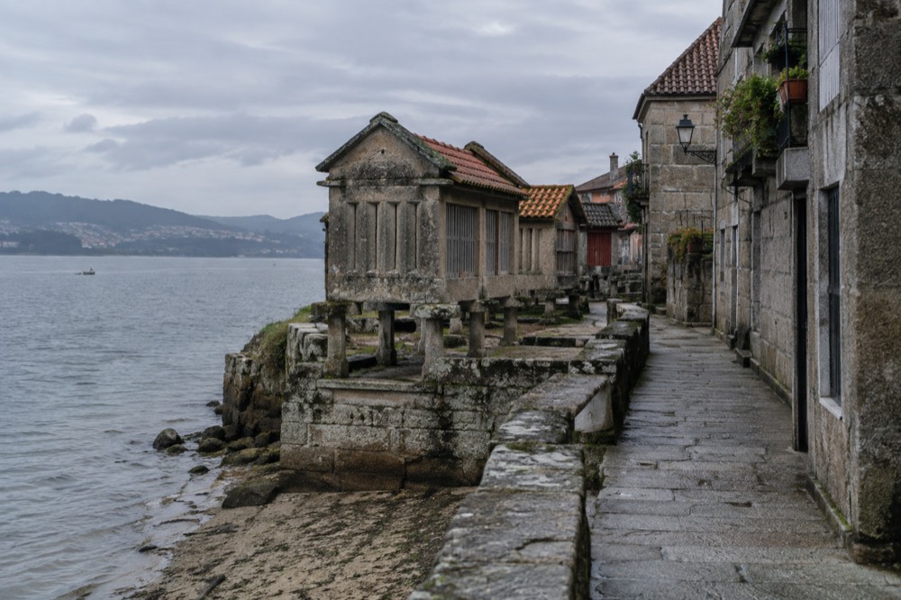 A narrow lane of stone houses above the Combarro waterfront, with laundry plants on balconies and the ria visible between buildings