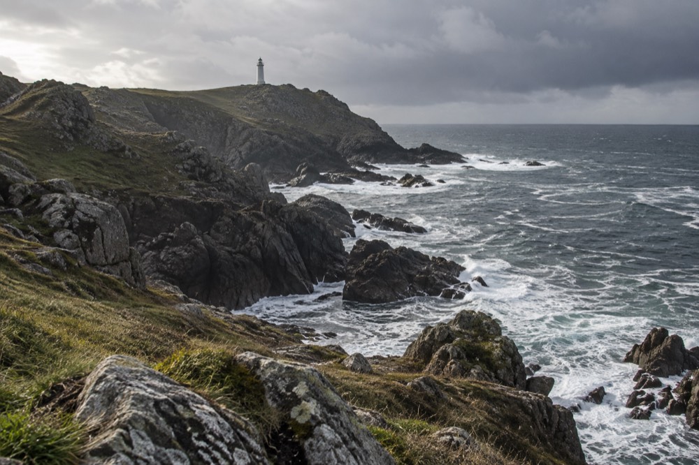 A white lighthouse tower standing above cliff grass at the edge of the ocean