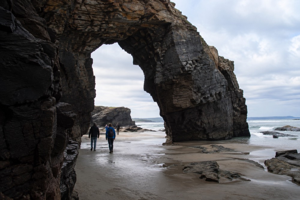 Walkers passing under a huge rock arch on a Galician beach at low tide