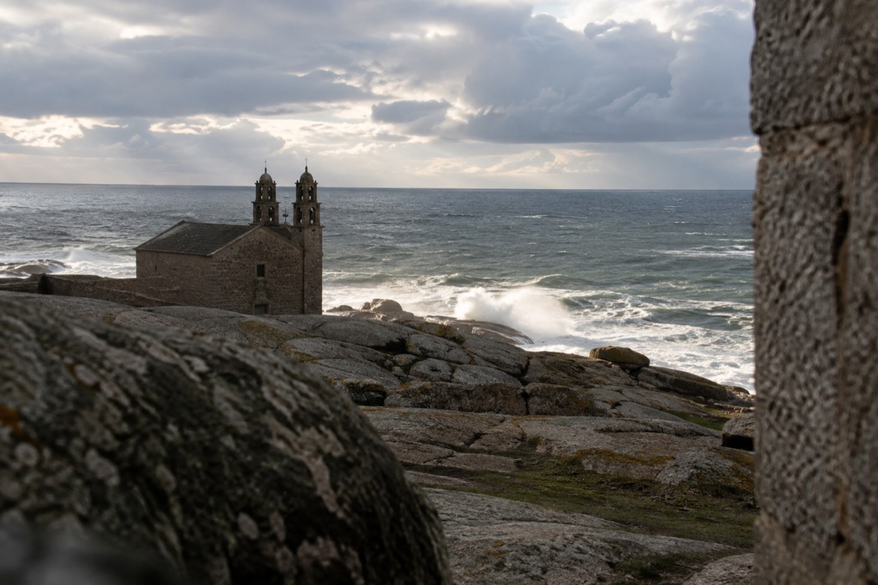 A stone sanctuary chapel standing on rocks beside the Atlantic Ocean in a small Galician coastal village