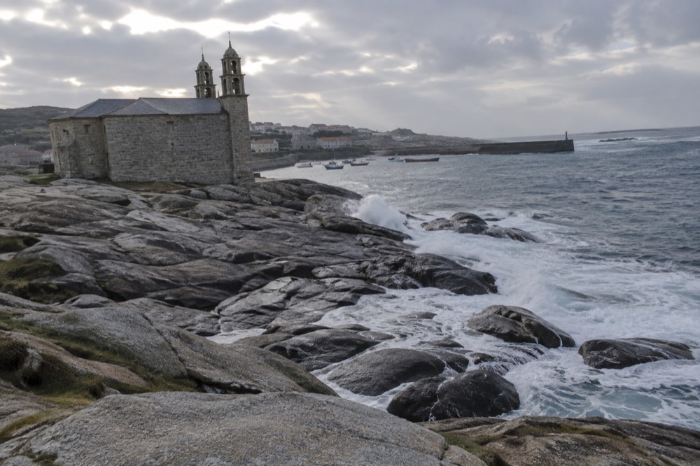 Fishing boats moored in a small harbour with grey stone buildings behind them under an overcast sky