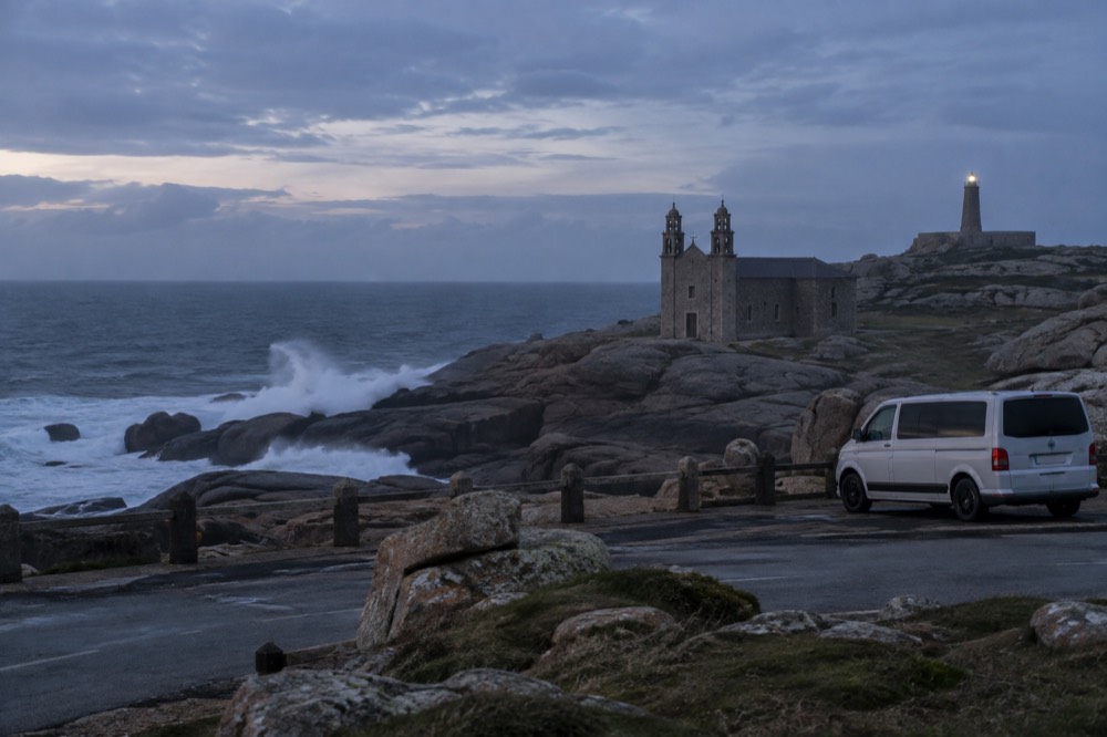 A tour van parked on a coastal road with a lighthouse and open ocean in the background