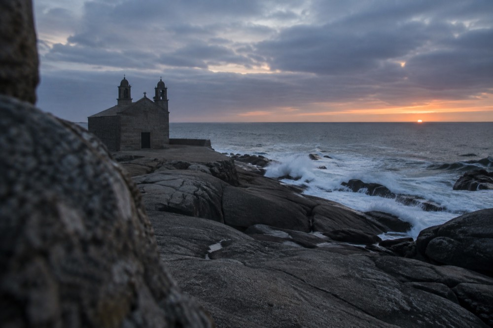 A granite chapel on a rocky point with waves crashing and a low sun over the Atlantic