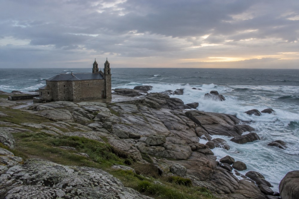 The stone church at Muxia's rocky point under a dusky sky, waves breaking over wave-worn granite at the Atlantic's edge