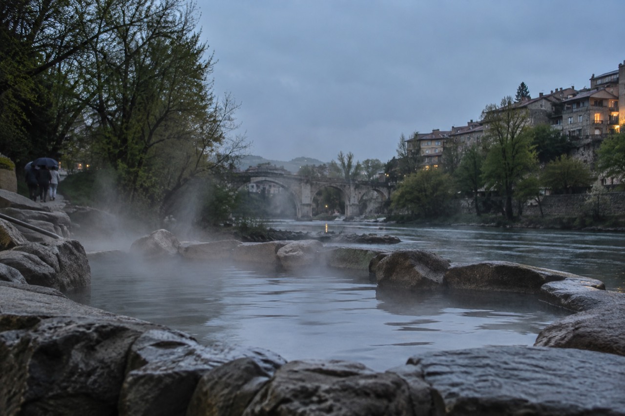 Steam rising from thermal spring water beside a river in a Galician provincial town with stone buildings on the far bank
