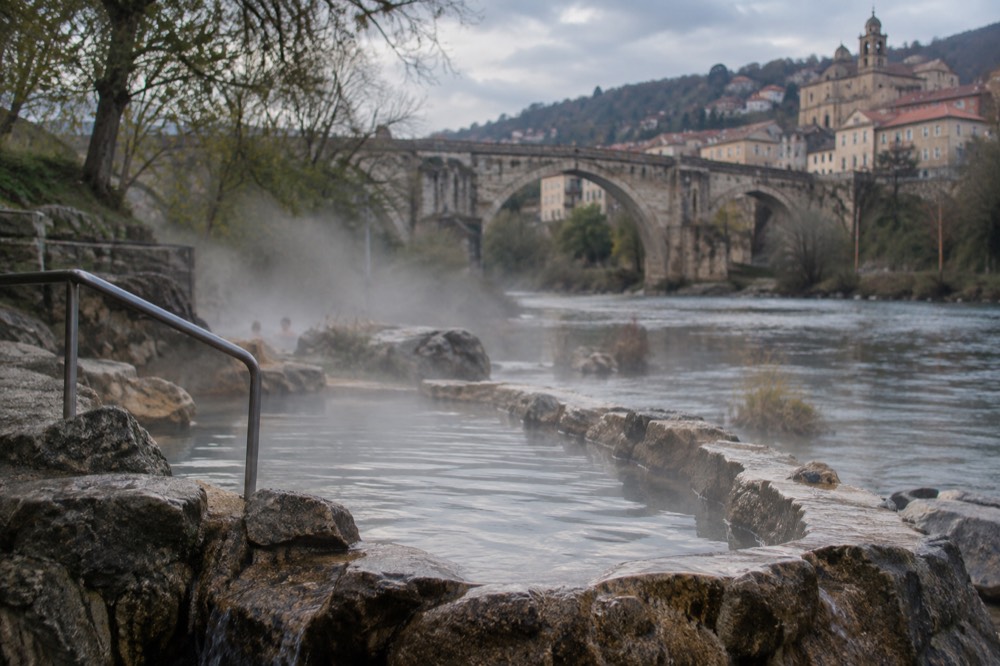 A stone Roman bridge arching over a river with a historic Galician town rising on the hillside beyond it