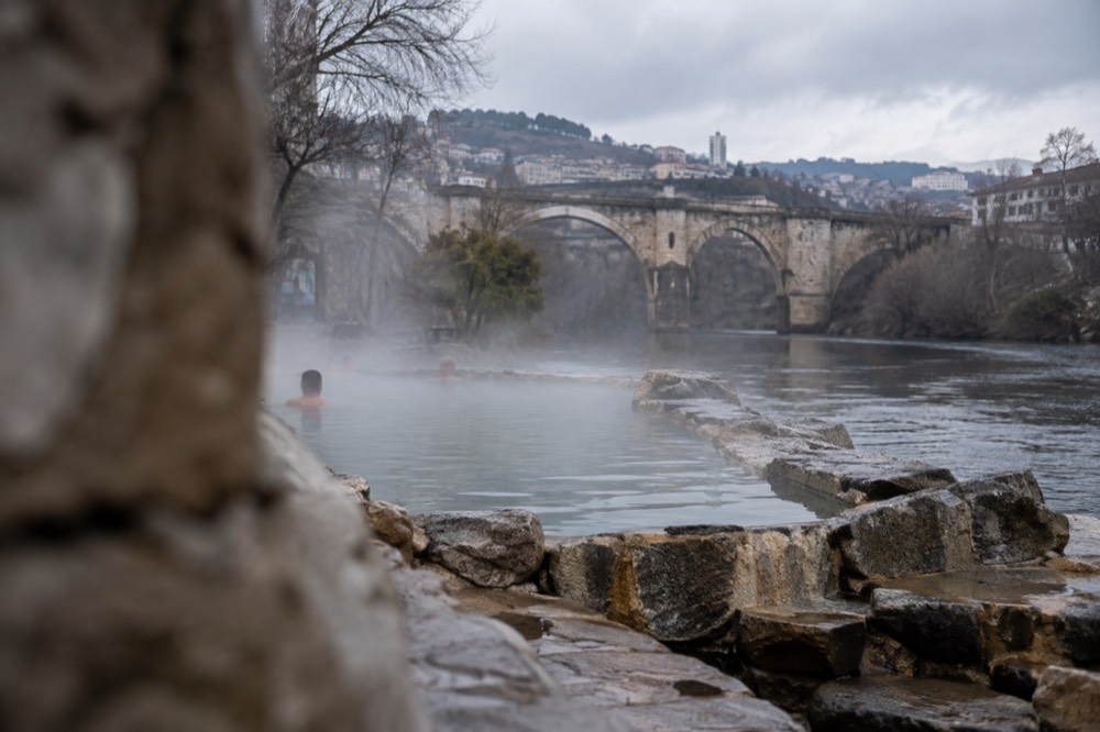 A riverside thermal pool at Ourense with steam rising off the water and a stone footbridge visible upstream