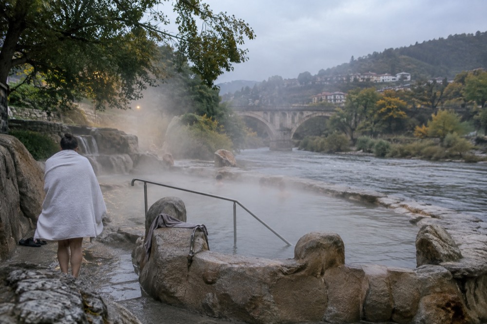 A visitor wrapped in a towel beside steaming outdoor thermal pools on a cool misty morning with green hills behind