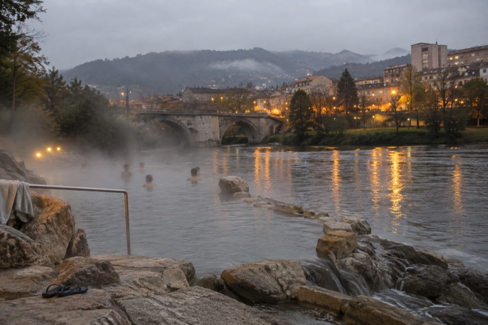 An evening view of a Galician provincial city with warm lights along a river, a stone bridge, and hills fading into mist beyond