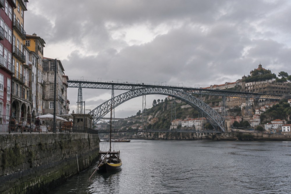 Tall narrow houses in faded render stacked above the Douro river in Porto's Ribeira district, terraced port-wine cellars on the far bank in evening light