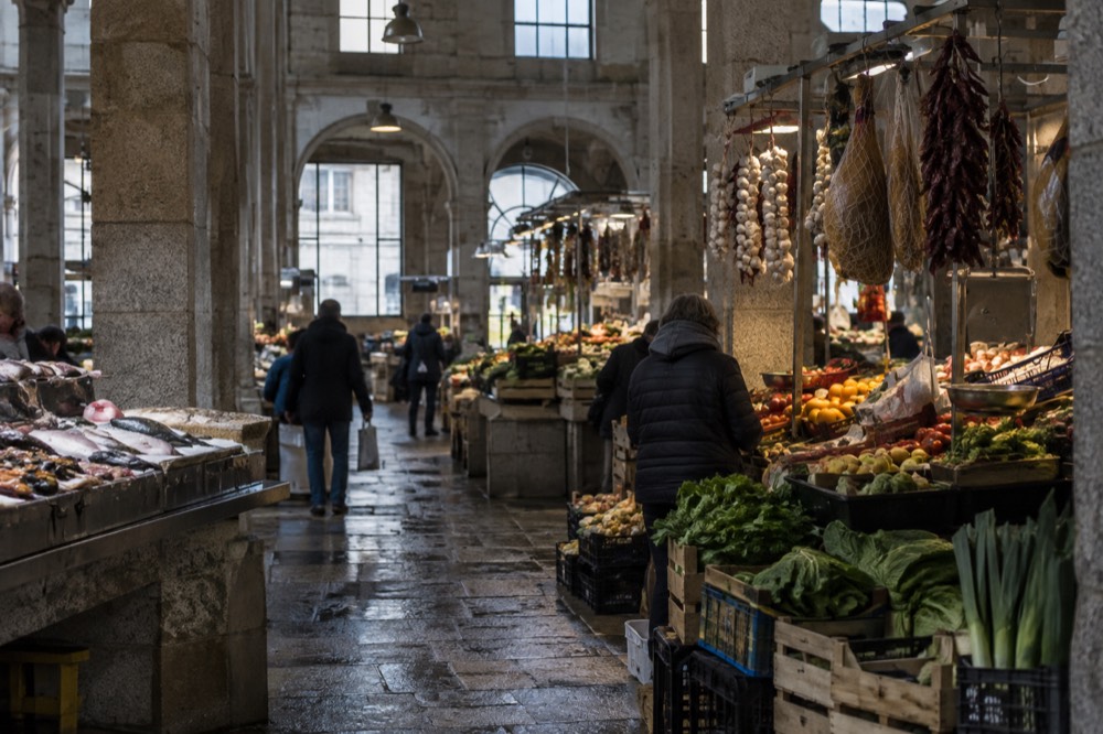 A market stall with stacked fruit crates and hanging cured goods inside a covered Galician market hall