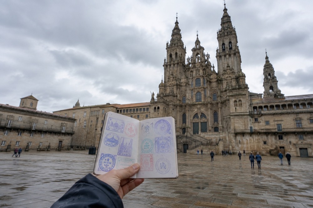A person holding an open pilgrim passport booklet filled with colorful ink stamps