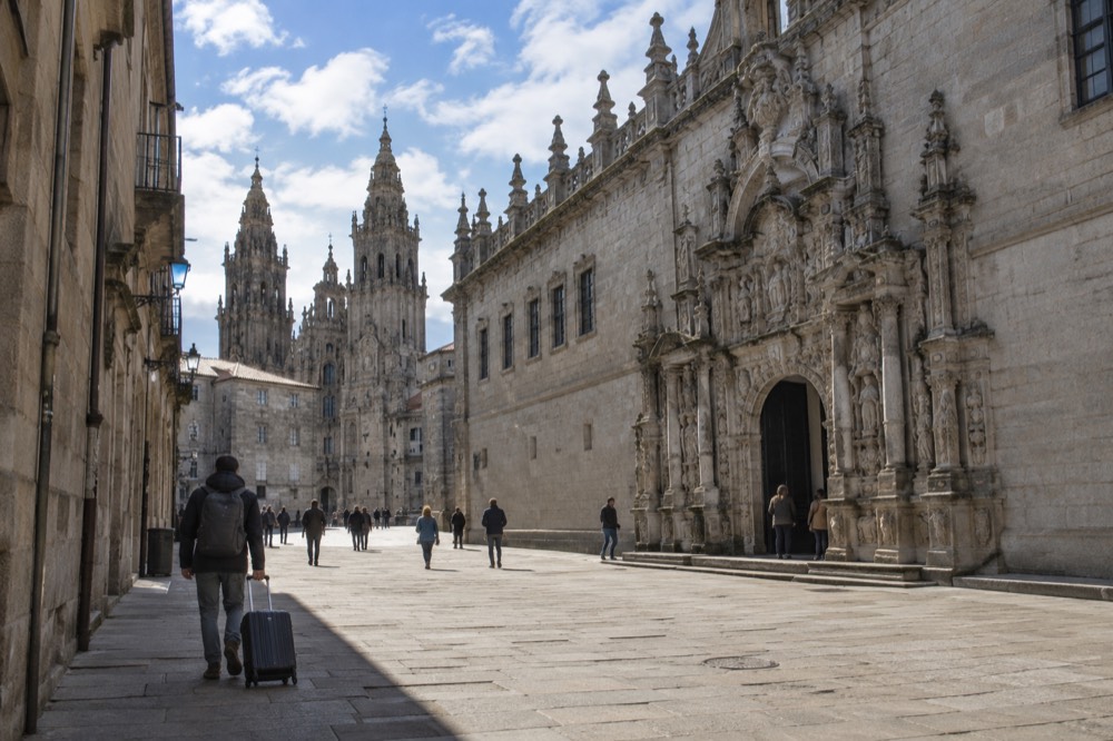Traveler with a suitcase walking up a narrow granite street toward the Cathedral towers in Santiago de Compostela