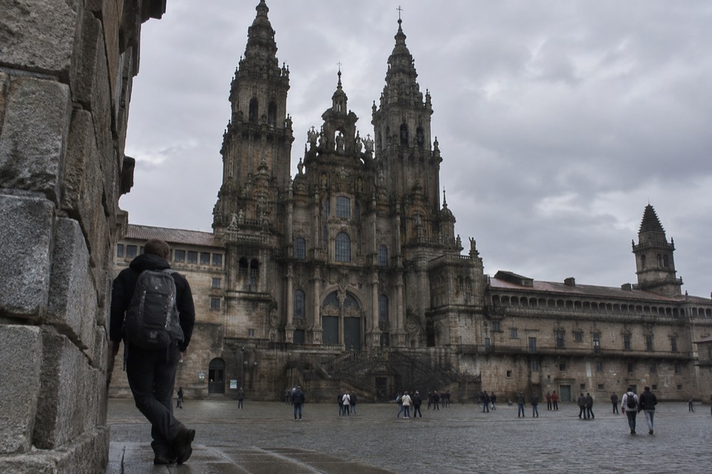 A pilgrim leaning a worn backpack against a stone wall beside a cathedral facade