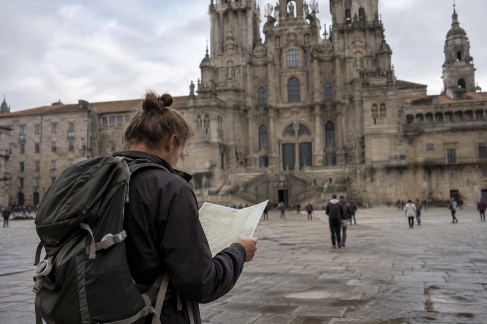 A pilgrim studying a folded paper map at a wooden table beside a walking backpack and hiking poles