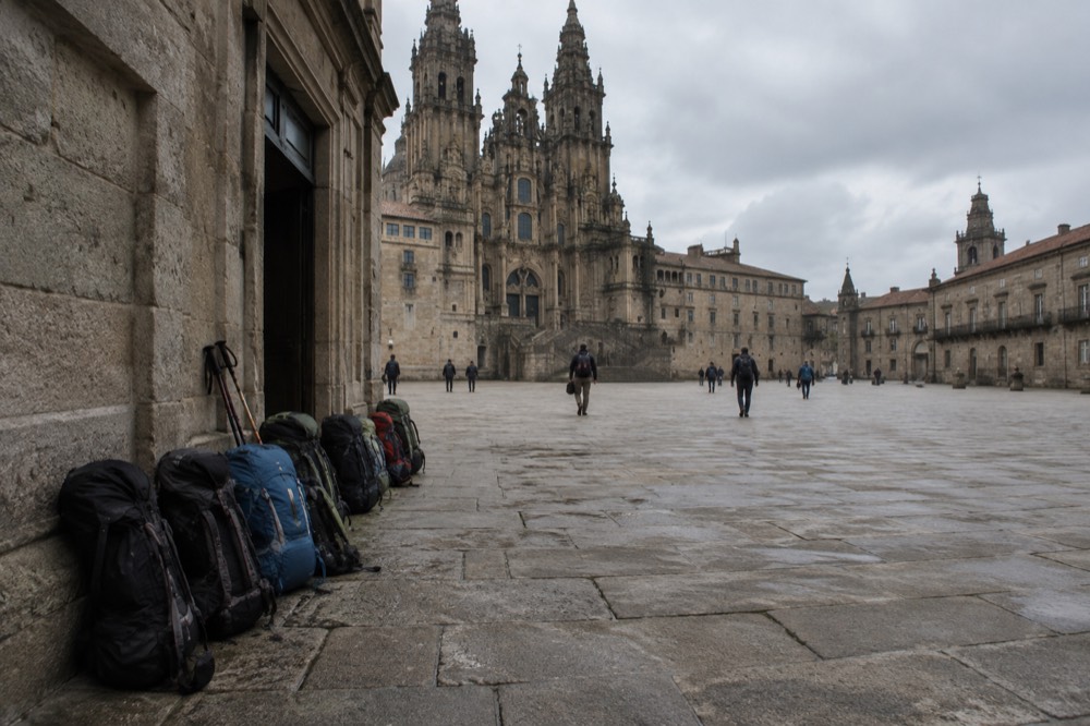 A row of pilgrim backpacks lined up outside a stone albergue entrance in the late afternoon