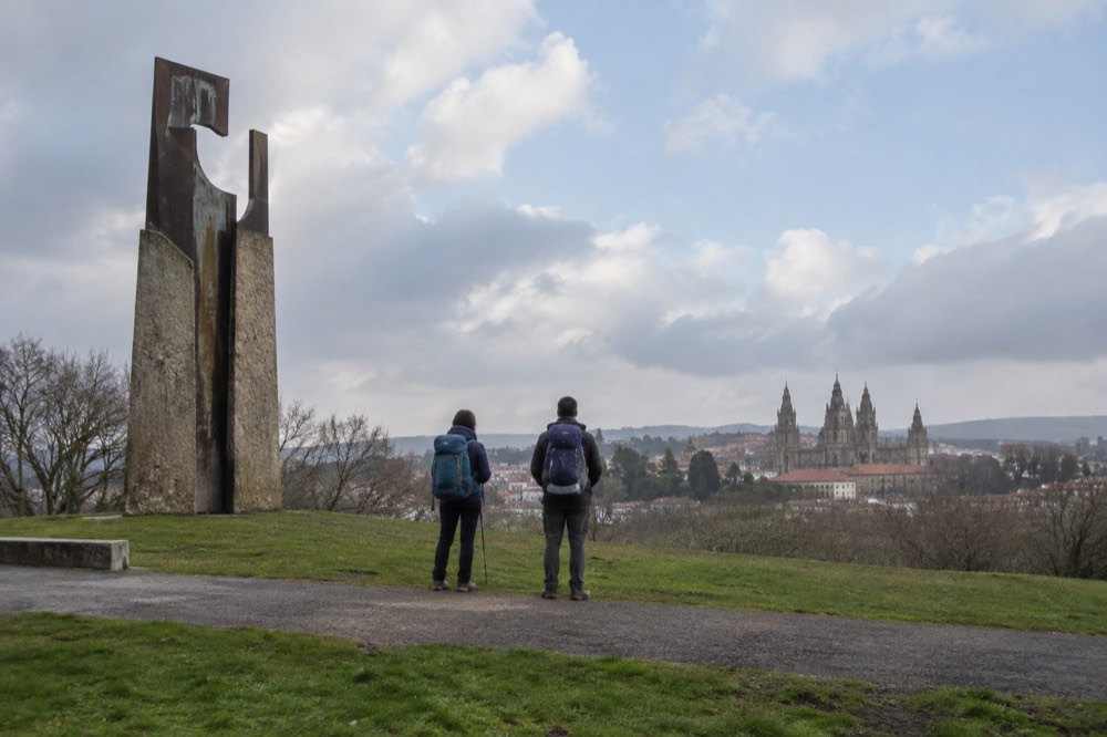 Pilgrims with backpacks standing on Monte do Gozo looking toward the distant towers of Santiago de Compostela Cathedral