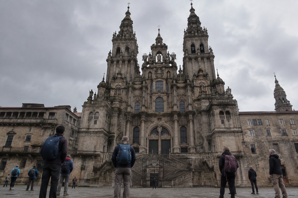 Pilgrims standing with backpacks in a wide granite square looking up at a cathedral facade