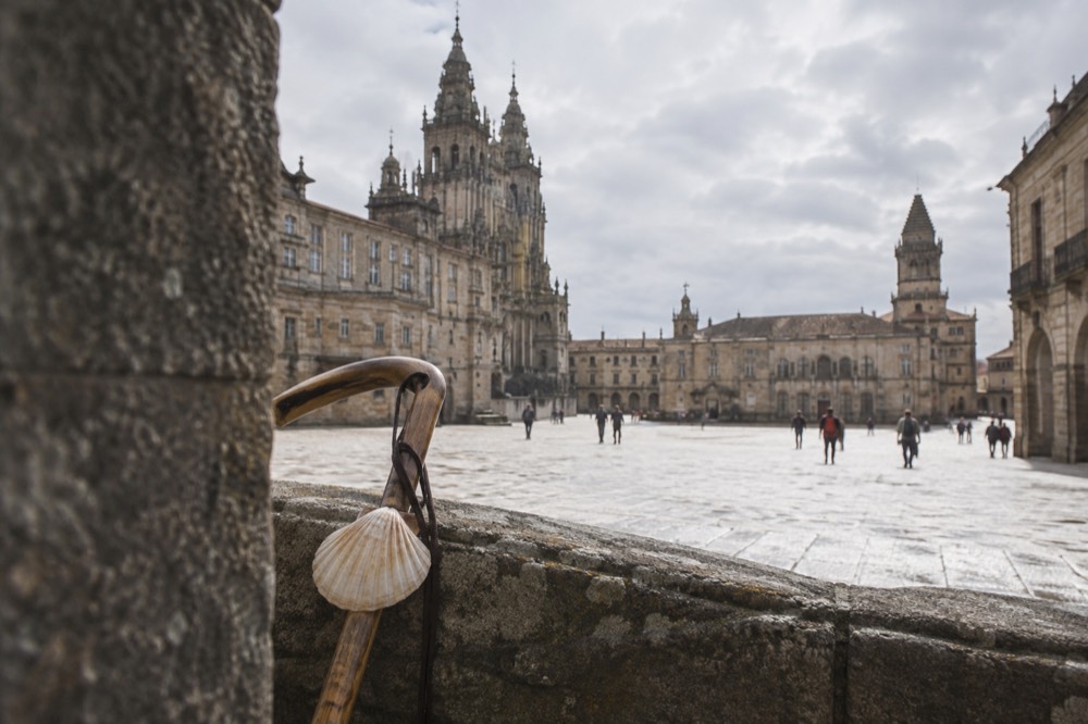 A scallop shell and walking stick resting on a stone parapet with a cathedral tower behind