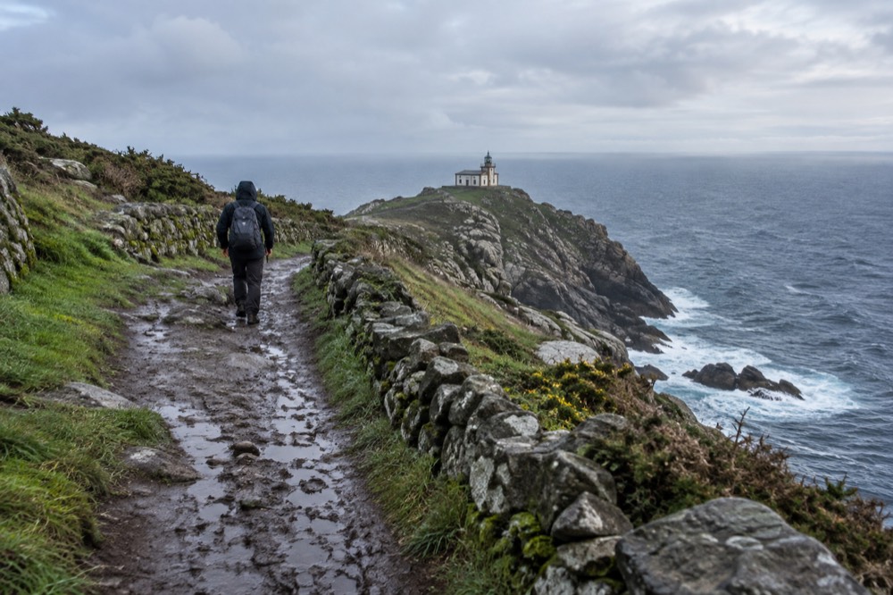 A pilgrim in a rain jacket walking a muddy rural track between stone walls in Galicia