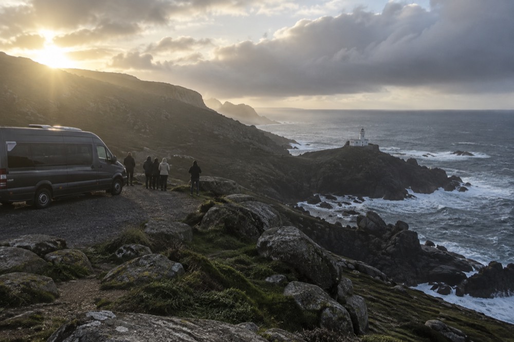 A coastal tour minibus parked at a viewpoint above a granite lighthouse on the Costa da Morte