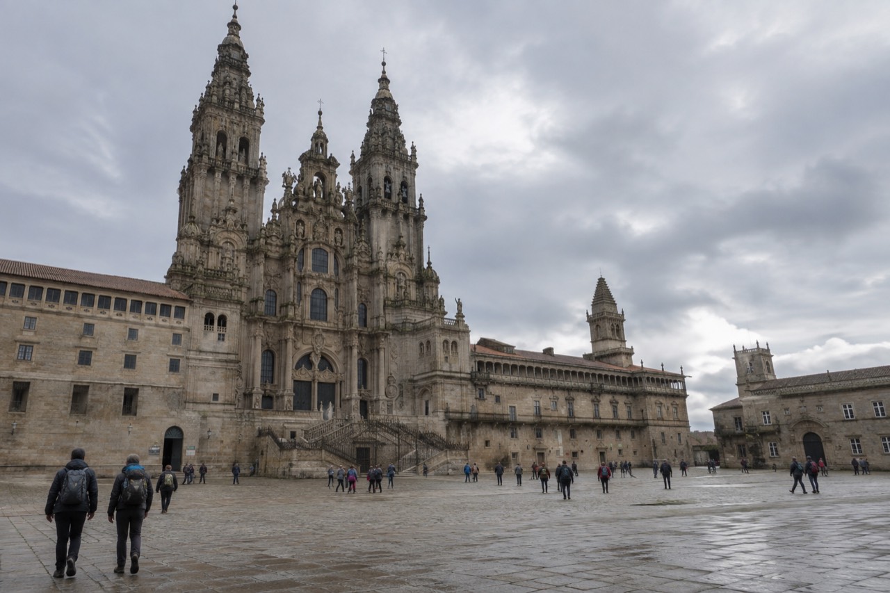 Pilgrims with backpacks walking a shaded rural path marked by a yellow arrow toward Santiago de Compostela