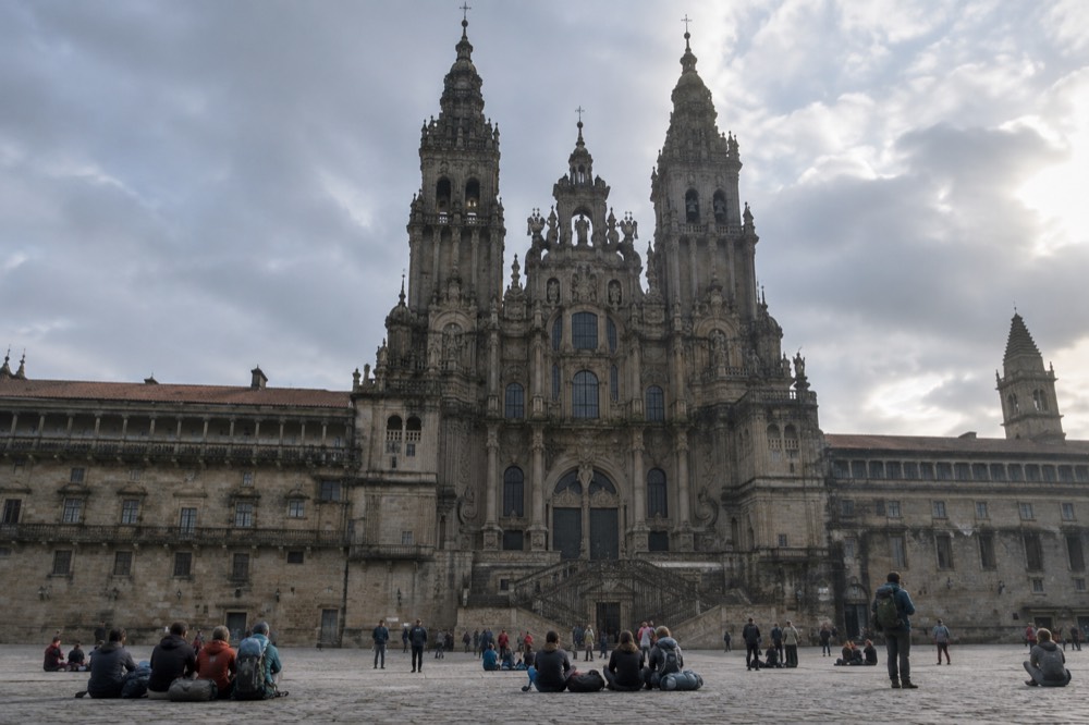 Pilgrims with backpacks resting on the stone paving of a large square in front of a baroque cathedral facade