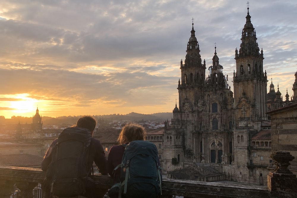 Two pilgrims leaning on a railing looking out over the rooftops and cathedral towers of Santiago de Compostela at sunset
