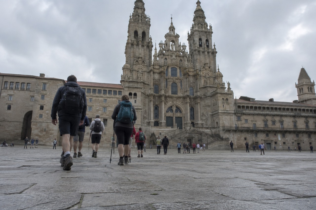 Pilgrims with small backpacks walking across a granite square toward a baroque cathedral facade