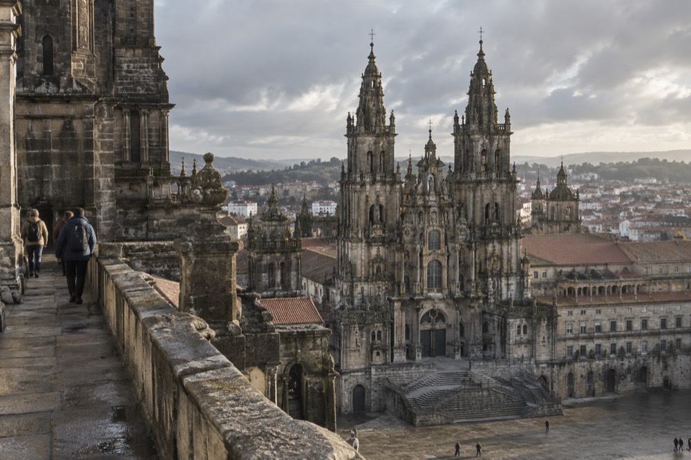 Walkers on a stone rooftop walkway beside a cathedral tower overlooking a city skyline