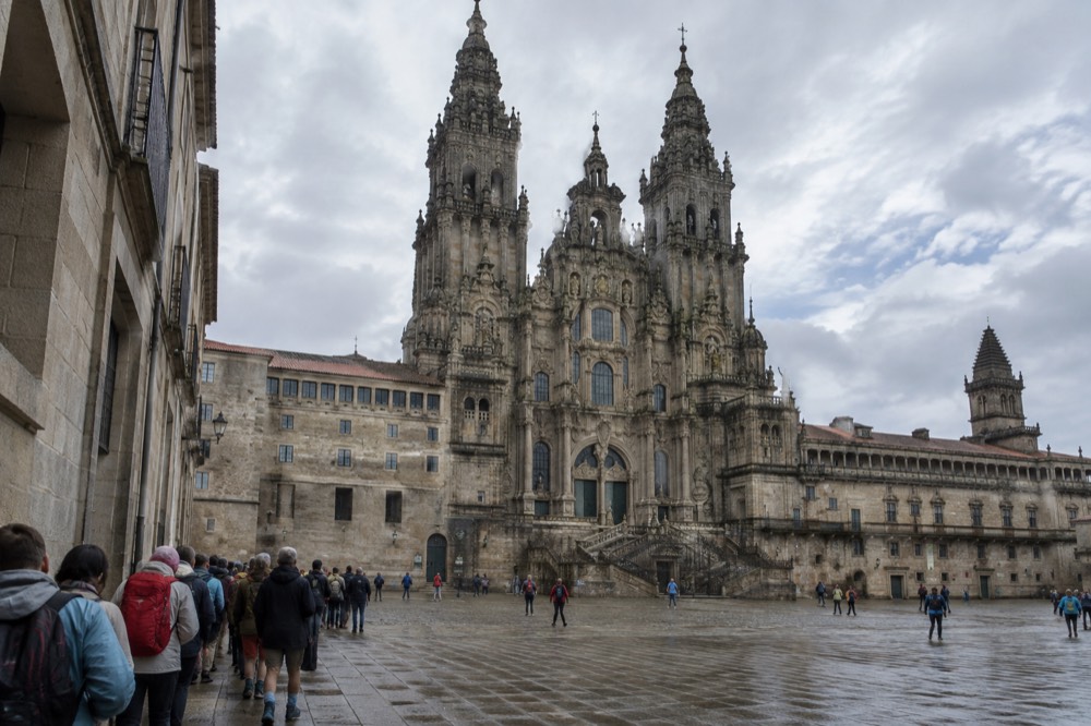 A queue of pilgrims with backpacks waiting outside the Pilgrim's Office on a narrow granite street in Santiago de Compostela