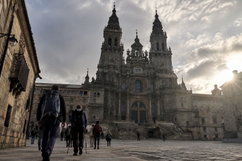 Pilgrims with backpacks walking into Santiago de Compostela past stone buildings toward the cathedral towers