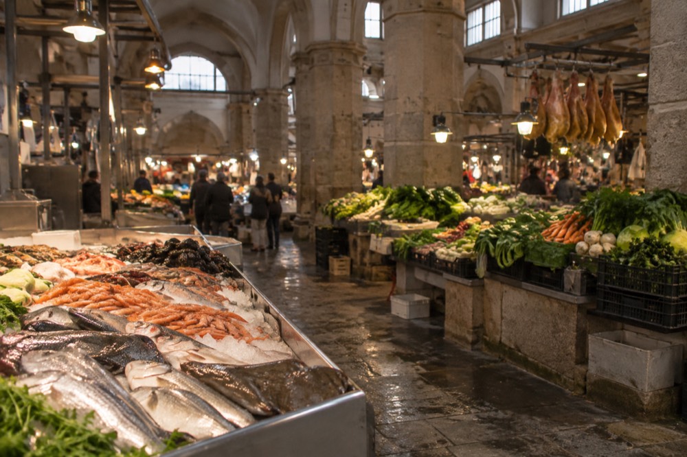 A busy indoor food market hall with stalls of fish, vegetables and hanging hams