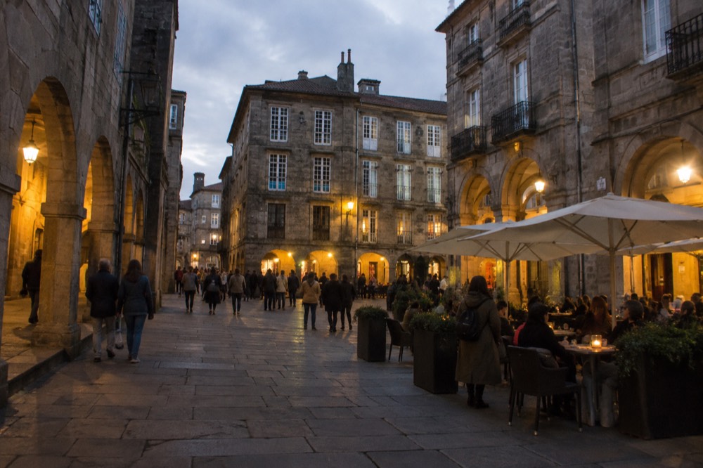 A lively stone square in the old town at dusk with crowds and café tables under warm lights