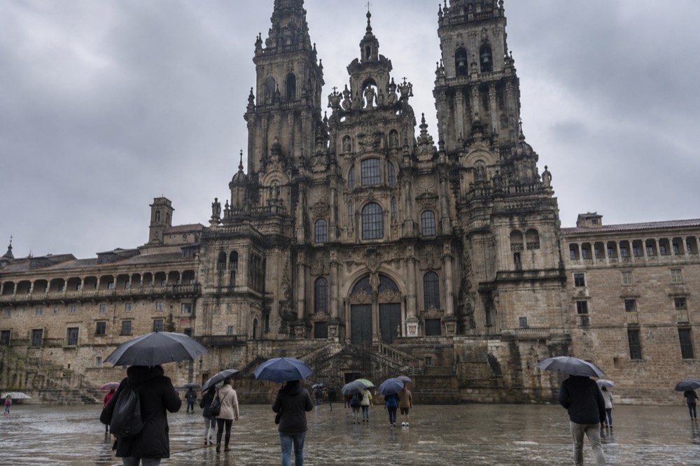 People with umbrellas crossing a wet granite square in front of a baroque cathedral facade